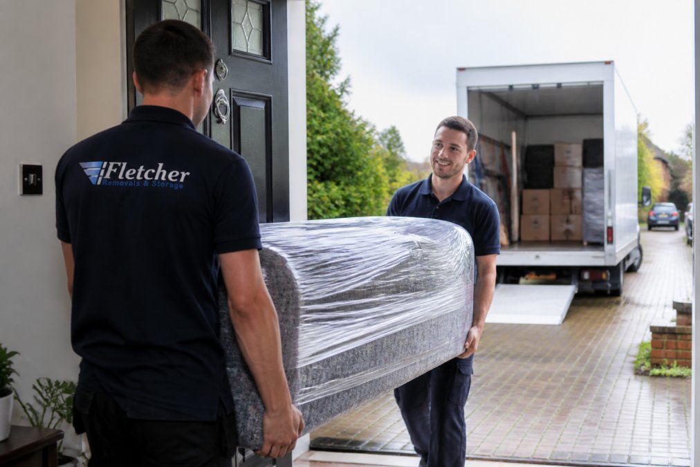 A uniformed removal team carefully carrying a wrapped sofa through the front door of a home, with a removals van parked on the driveway behind them