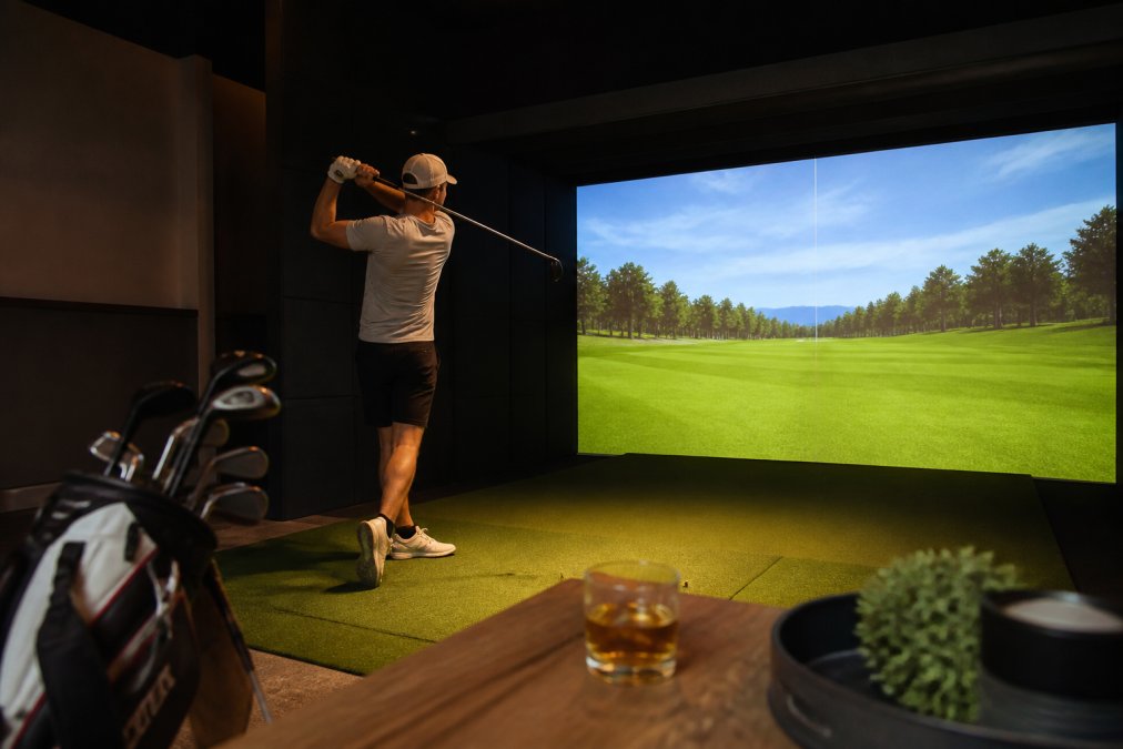 Golfer mid-swing in private simulator bay with Full Swing screen visible in background displaying ball flight trajectory, shot data, and spin rate metrics