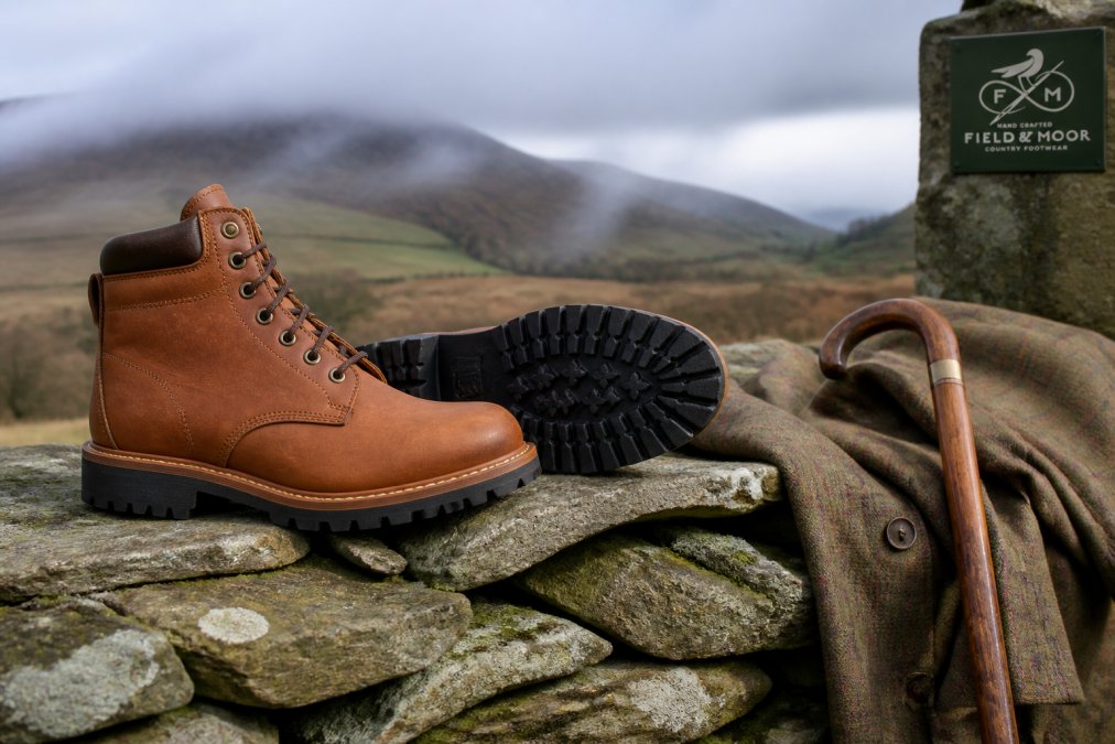 A pair of handcrafted tan leather country boots resting on a stone wall in a misty countryside setting, with a walking stick and tweed jacket nearby