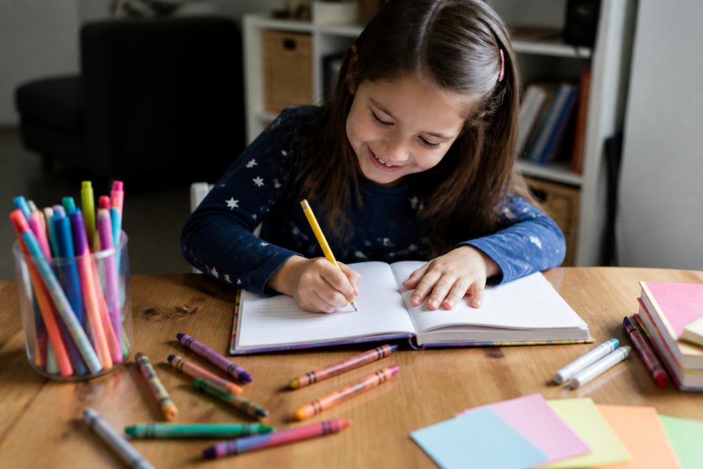 Young elementary student writing in a colorful journal at a desk with crayons and markers nearby, smiling while engaged in creative journaling