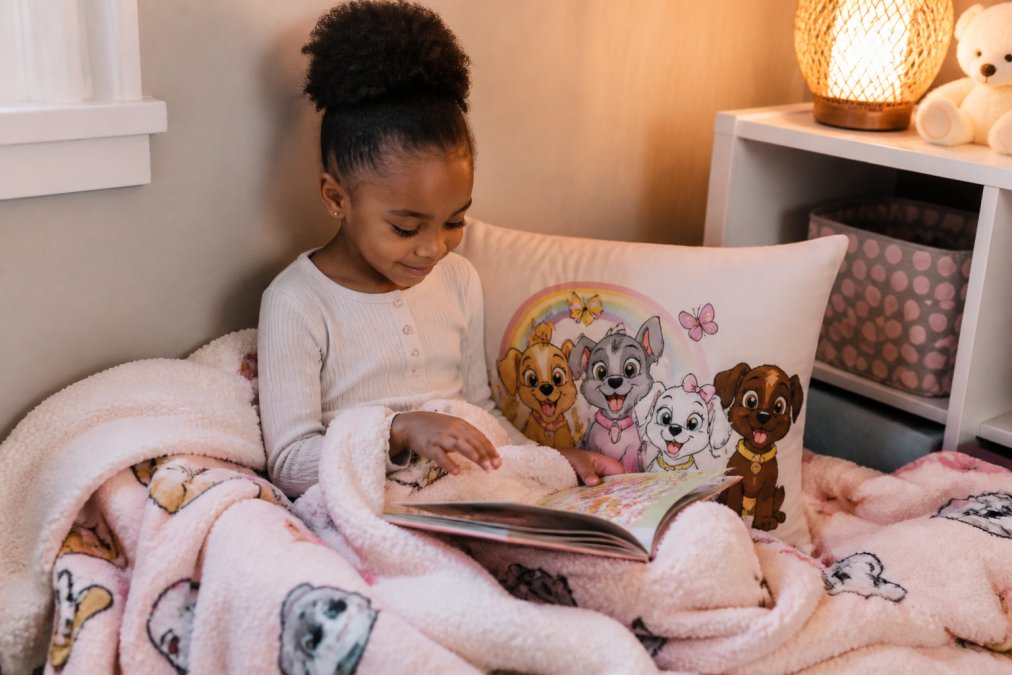 A young child sitting in a cozy reading nook corner with a plush sherpa blanket, a decorative pillow featuring cartoon puppies, and an open storybook on their lap, warm soft lighting in the background