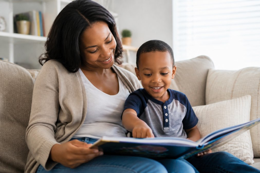Parent and young child sitting together on a cozy couch reading a colorful picture book, child pointing at illustrations with interest and smiling