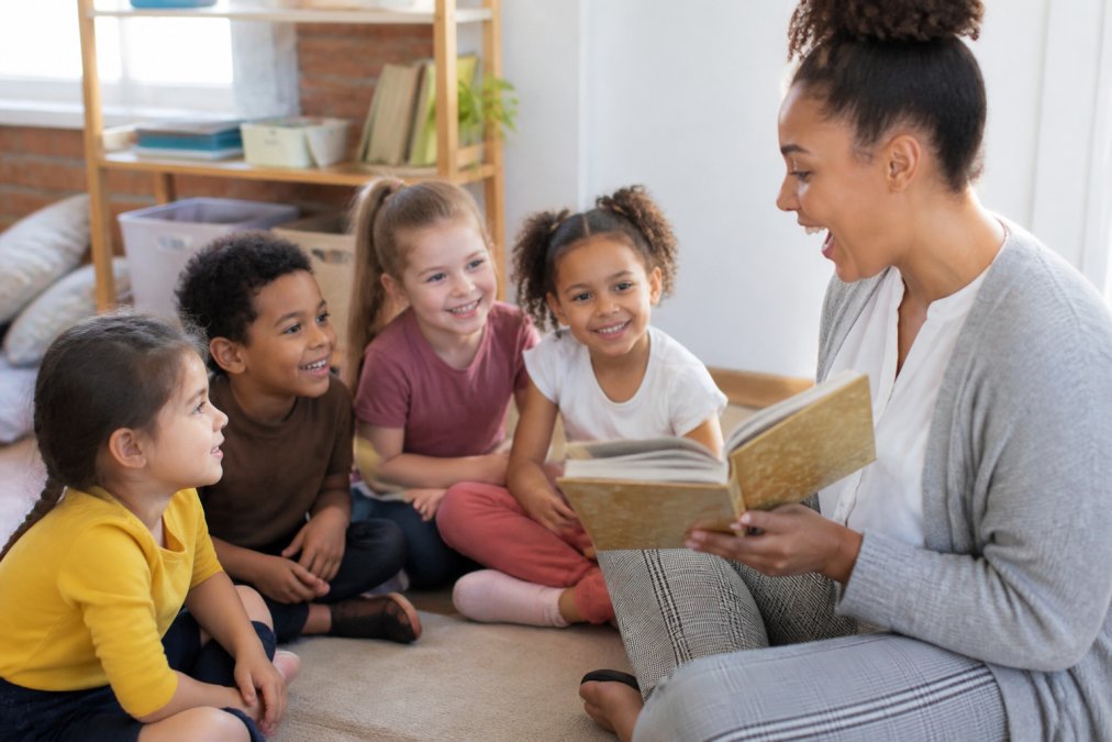 Small group of children sitting in a circle during a read-aloud session, engaged and smiling as an adult reads from a book with animated expression