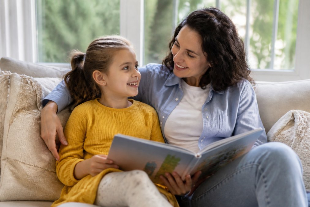 Parent and child sitting together on a comfortable couch by a bright window, child smiling while holding a colorful picture book, both engaged and relaxed