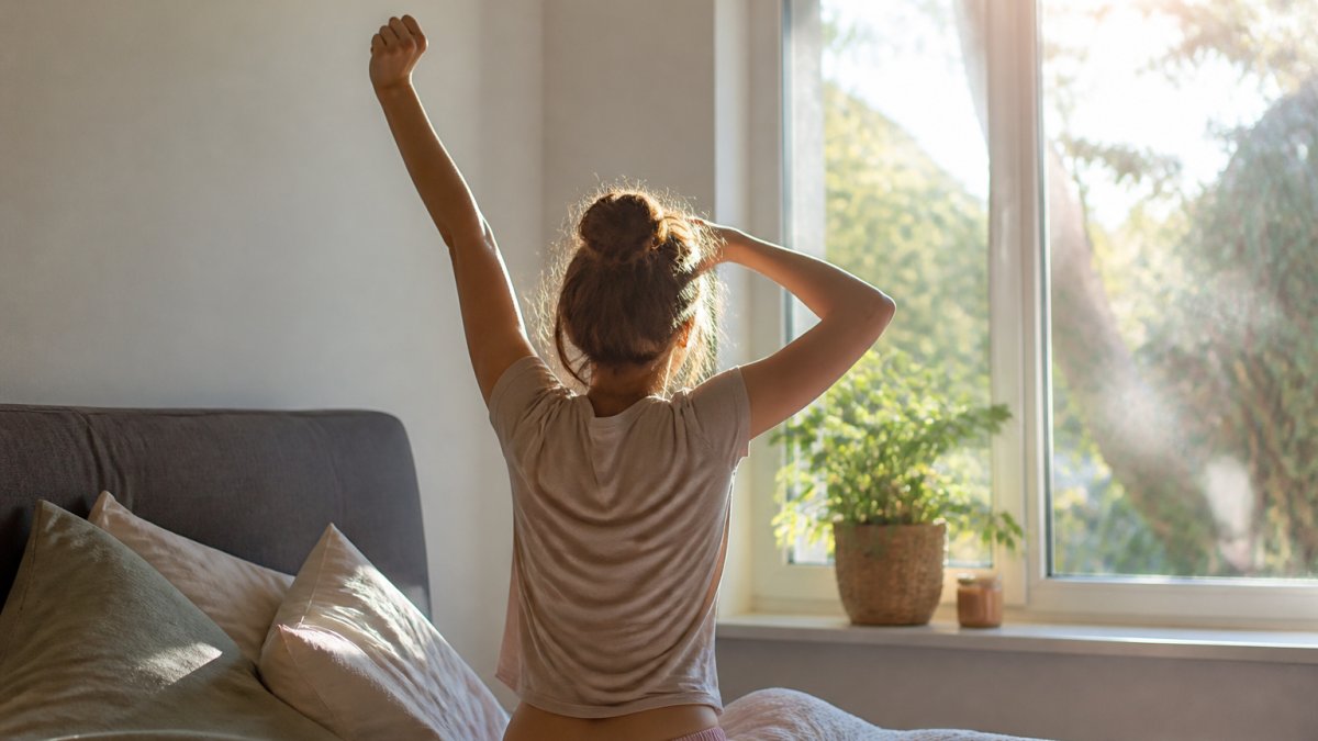 Person stretching in morning sunlight at window, showing fatigue and low energy body language, natural home setting