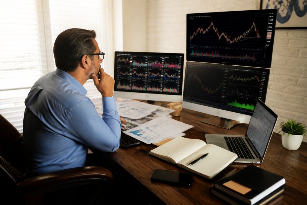 Professional investor reviewing multiple computer screens displaying financial dashboards and stock charts in a well-lit home office, with notebooks and financial documents on the desk