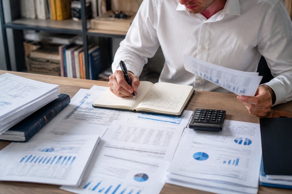 Professional analyst reviewing printed financial statements and making detailed notes with a calculator and notebook on a wooden desk, with charts and company reports spread across the surface