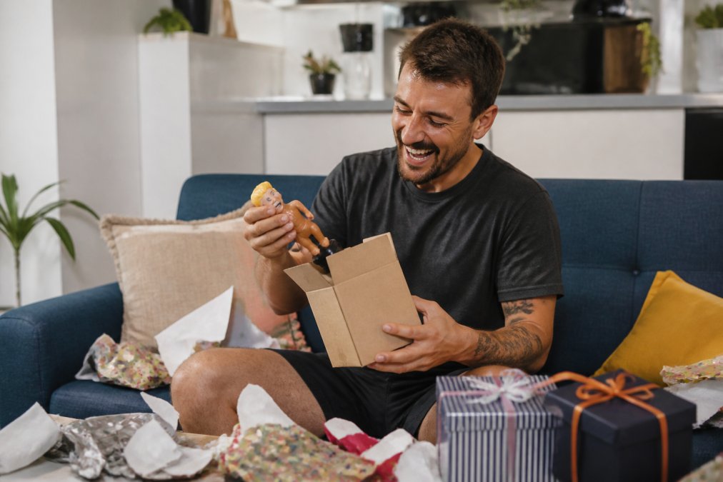 A man laughing whilst unwrapping funny gifts for men in a bright, modern living room with scattered paper.