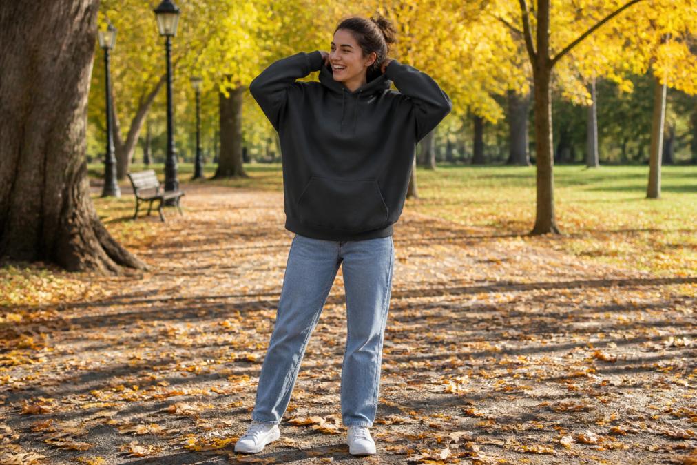 A person laughing while adjusting the hood of an oversized graphic hoodie, wearing jeans and sneakers, standing in a sunlit park with autumn leaves around