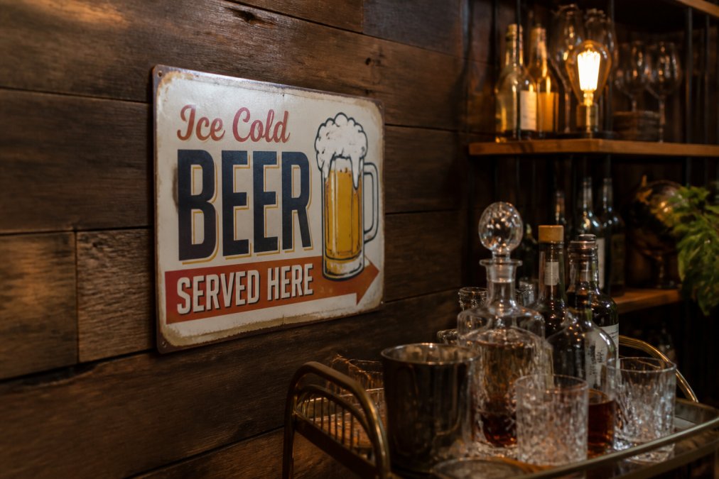 Vintage aluminum beer sign mounted on a dark wood wall above a bar cart with glassware and bottles in warm lighting.