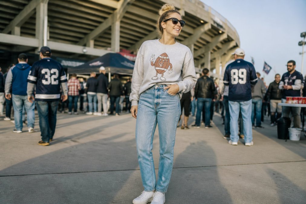 Person wearing a funny sports graphic tee layered under a crewneck sweatshirt, paired with high-waisted jeans and white sneakers, standing at a stadium entrance with other fans in the background and natural afternoon lighting