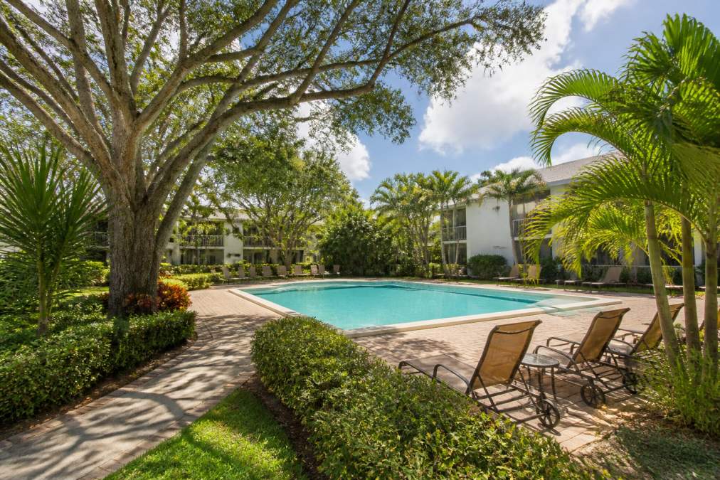 Resort-style heated pool surrounded by lush tropical landscaping, mature trees creating natural shade, and garden pathways with residents relaxing on lounge chairs in afternoon sunlight