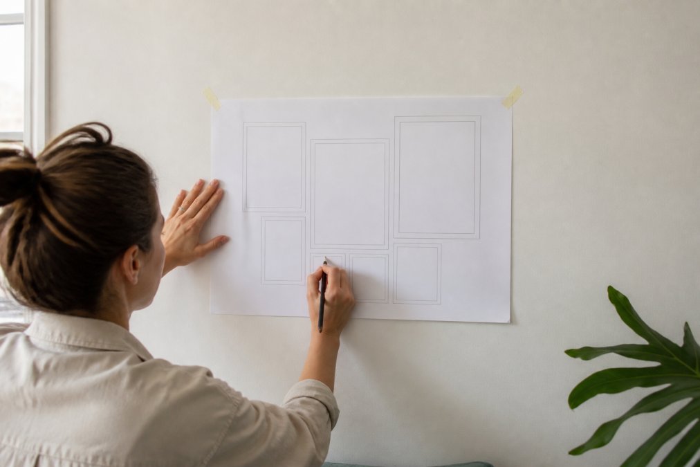 Hands holding a paper template against a blank white wall with a pencil marking positions, showing measurements and layout planning for a gallery wall installation in a small bright room