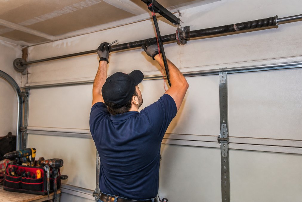 A professional technician in work clothes inspecting a residential garage door's torsion spring and track system with tools nearby