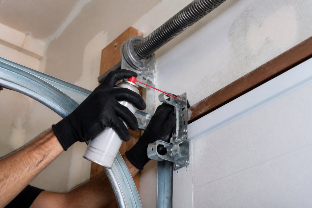 Close-up of a technician's gloved hands applying lubricant spray to garage door rollers and hinges, with the metal track and coiled torsion spring visible in the background of a residential garage