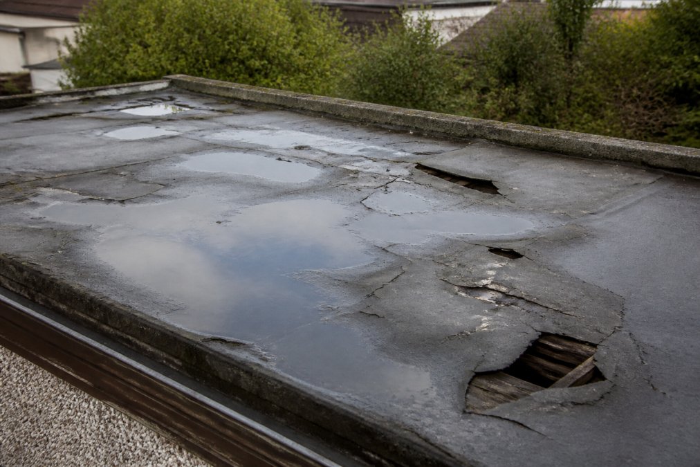 Damaged garage roof showing visible pooling water, cracks in rubber membrane, and deteriorated felt with daylight visible through gaps in the structure