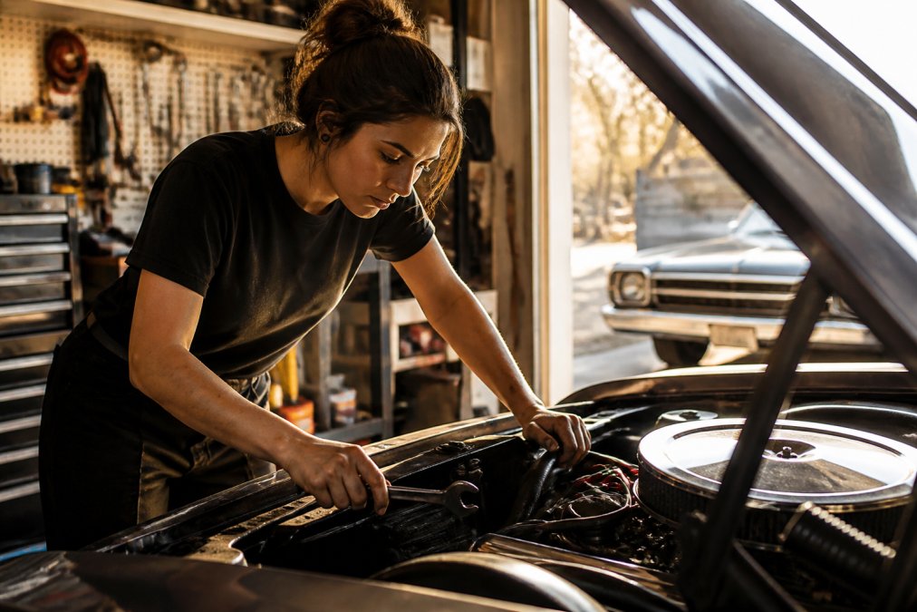 Female mechanic in a black graphic tee using a wrench to repair a classic car engine in a sunny garage