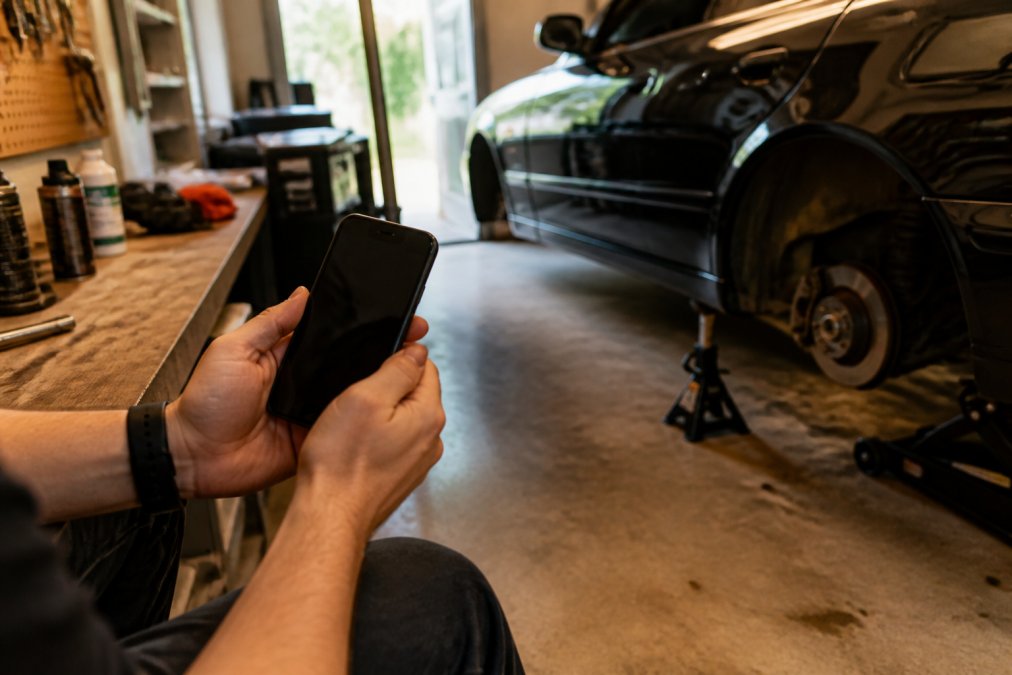 A person's hands holding a smartphone displaying a car maintenance app interface, with a project car on jack stands visible in a home garage in the background, warm workbench lighting