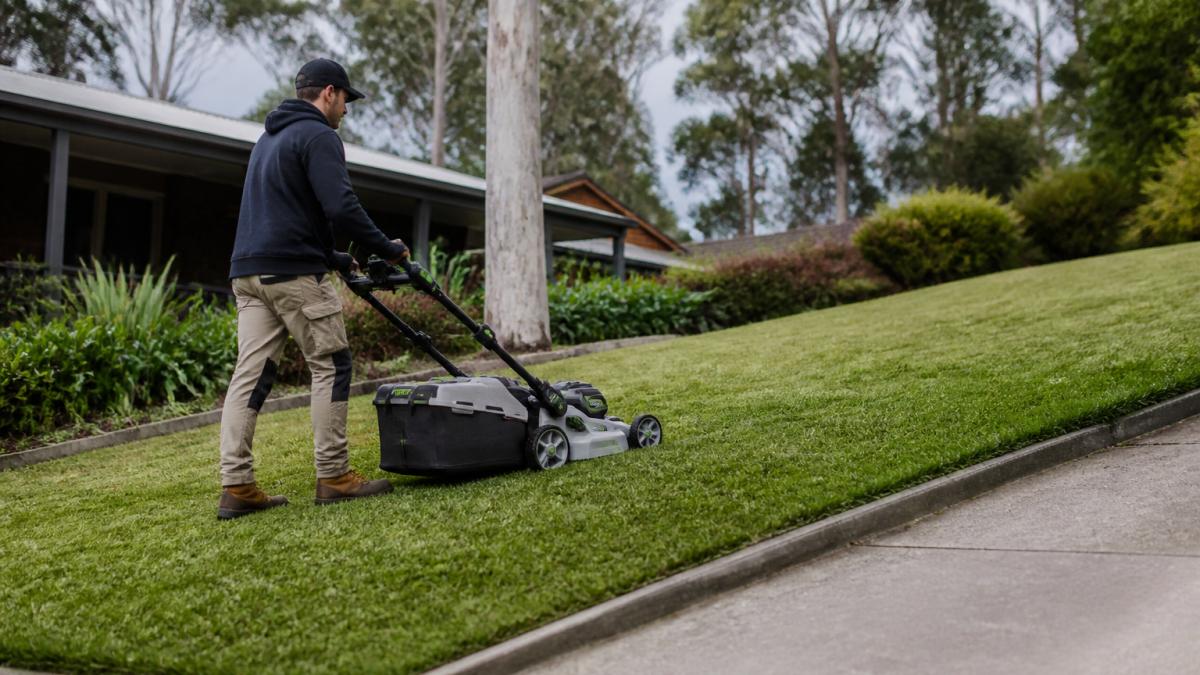 A person in work clothes pushing a battery-powered lawn mower across a sloping suburban lawn, with a neatly edged path visible and gum trees in the background