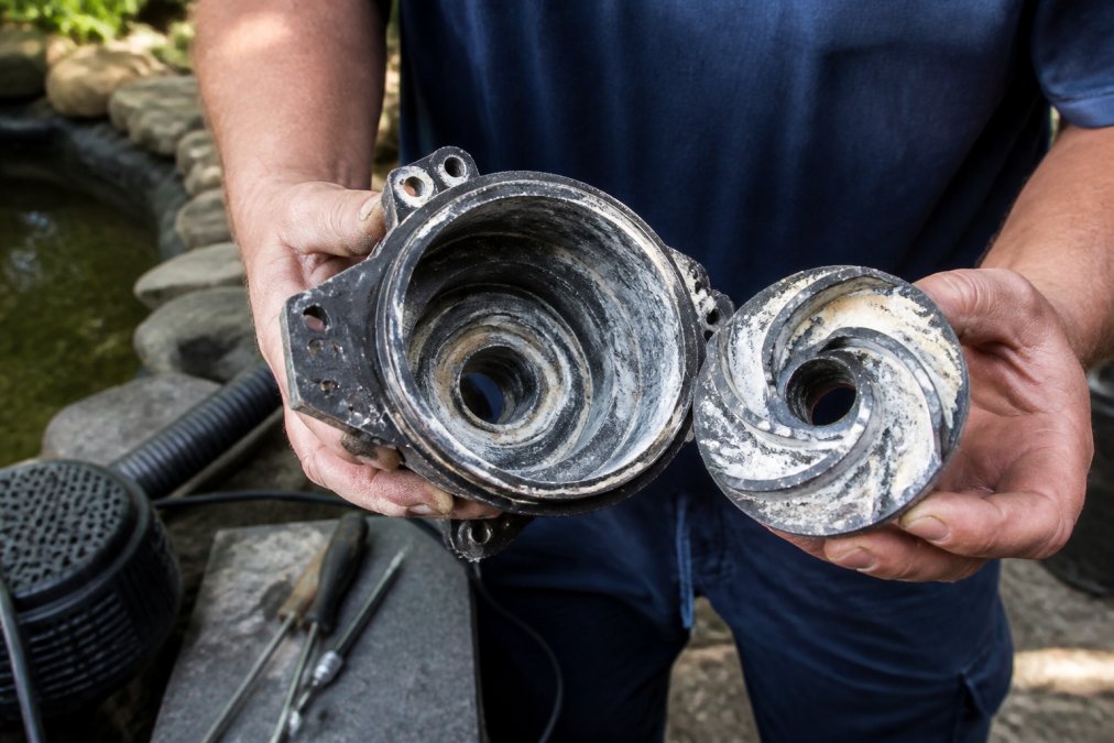 Close-up of a pond pump impeller and housing assembly held in hands, showing visible white calcium scale buildup and mineral deposits on the metal surfaces, with inspection tools visible in soft natural lighting