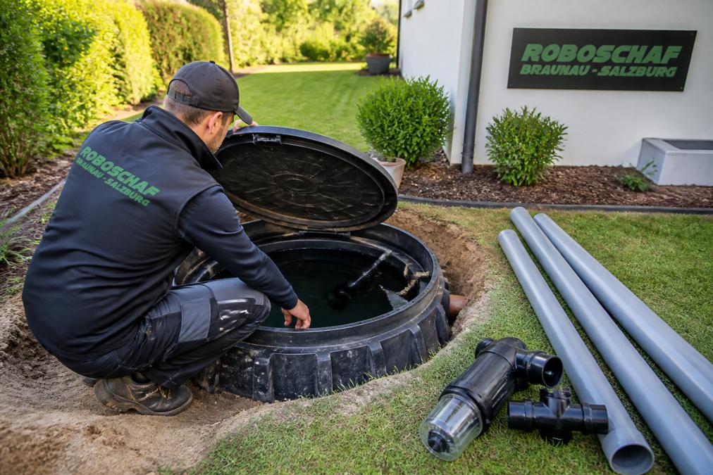Person in Arbeitskleidung prüft eine unterirdische Regenwasserzisterne im Garten, daneben liegen Rohre und ein Filter für die Bewässerungsanlage, Tageslicht
