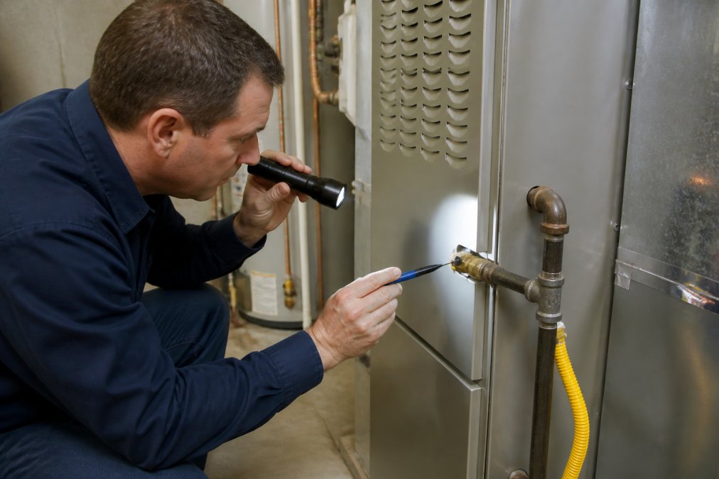 Homeowner using a flashlight to inspect furnace air intake and gas piping connections, pointing to visible components with a pen