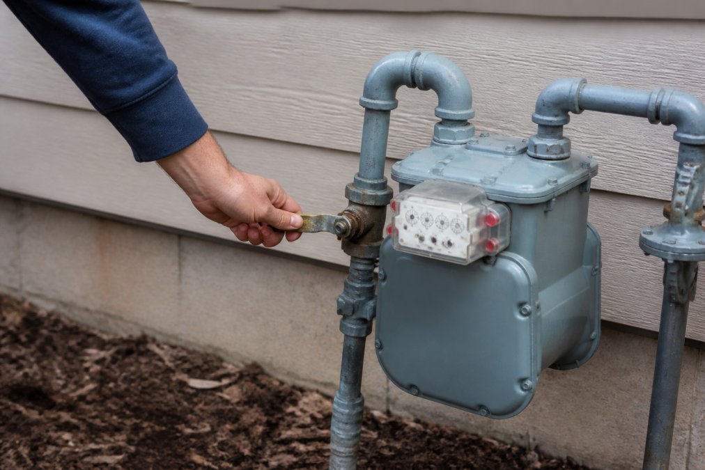 Hand reaching toward gas meter and shut-off valve on home exterior, showing valve handle and meter clearly