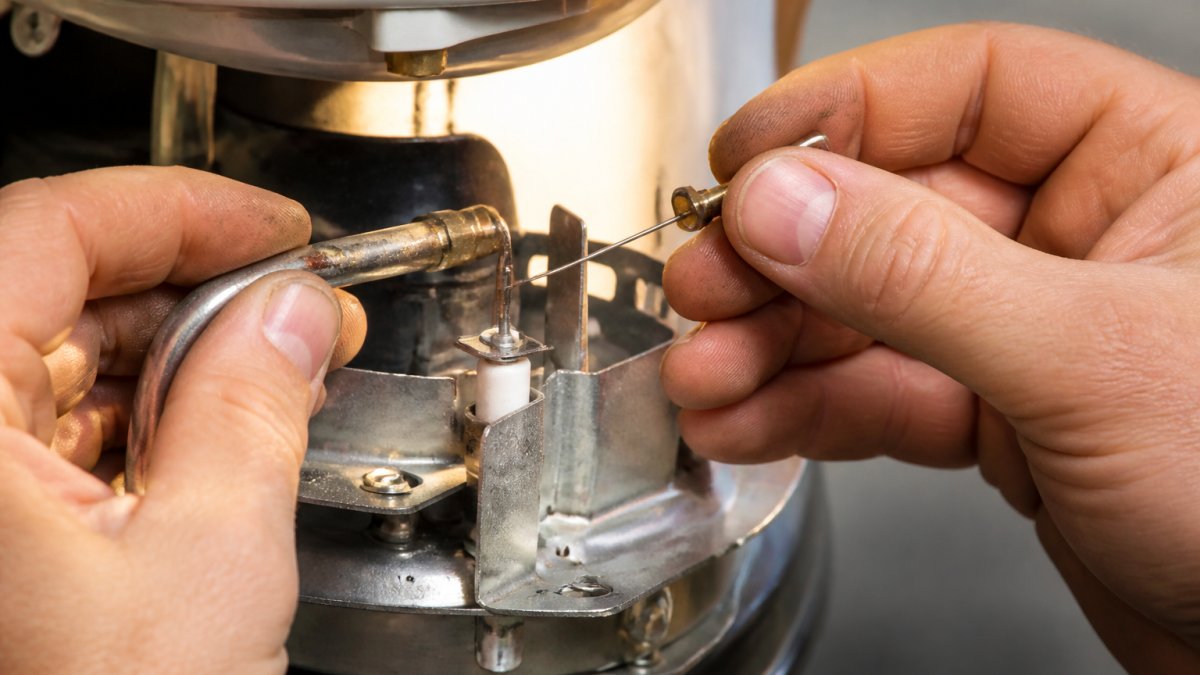 Professional technician's hands using a small needle or wire to carefully clean the pilot light orifice on a water heater burner assembly, showing precise detail work under bright task lighting