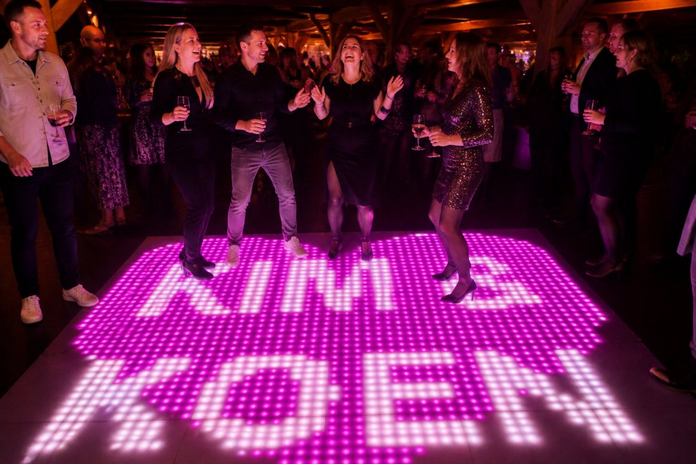 Groep feestgangers dansend op een verlichte dansvloer met kleurrijke LED-tegels, met energieke bewegingen en vrolijke gezichten terwijl het gekleurde licht hun gezichten verlicht