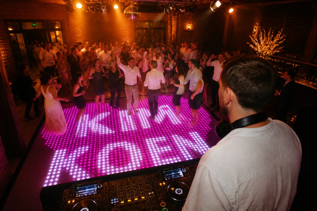 DJ achter de draaitafel terwijl hij het publiek op de dansvloer observeert, met gasten die dansen en genieten, warm licht boven de dansvloer