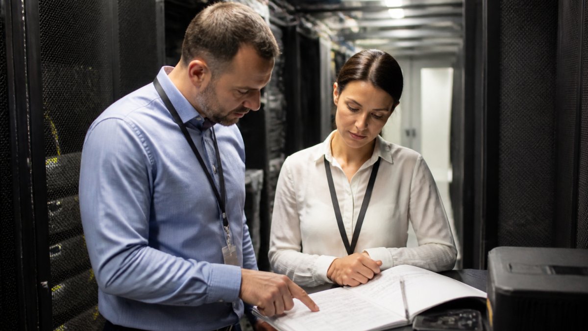 A database administrator and a QA lead reviewing masking configuration on a monitor in a secure server room, printed audit report on the desk beside them