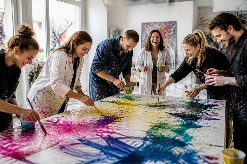 Gruppe von Erwachsenen malt gemeinsam an großflächigen Kunstwerken in bunten Farben, lachend und konzentriert mit kreativer Energie in einem hellen, modernen Studio