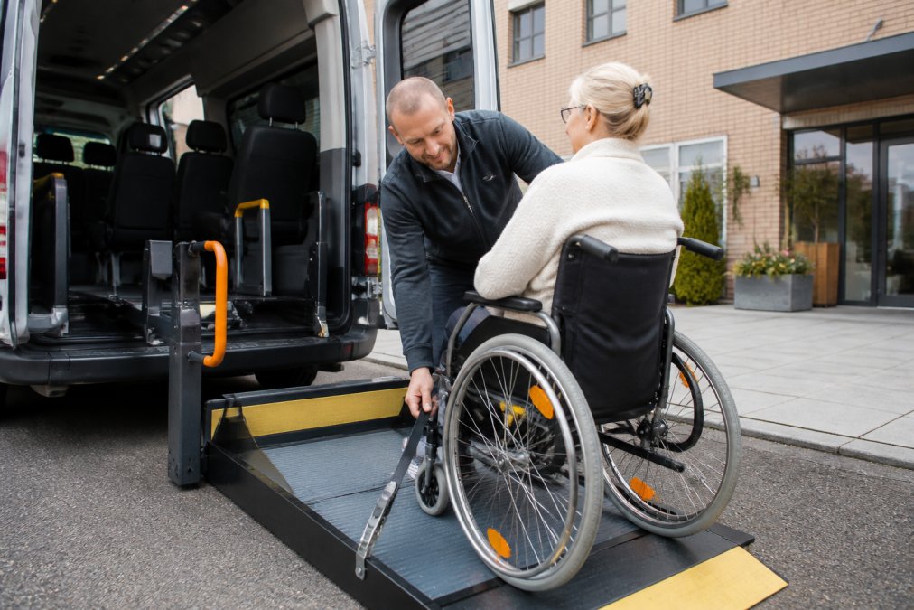 Gecertificeerd rolstoelvervoer: chauffeur helpt passagier veilig in een aangepast voertuig bij een kliniek.