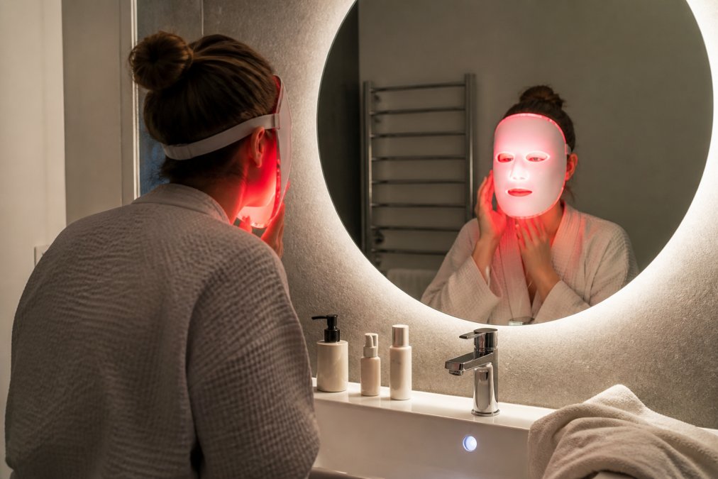 Een persoon die 's avonds een gezichtsbehandeling thuis uitvoert met een PDT lichttherapiemasker op een goed verlichte badkamerspiegel, met een paar verzorgingsproducten op de wastafel en een handdoek over de rand