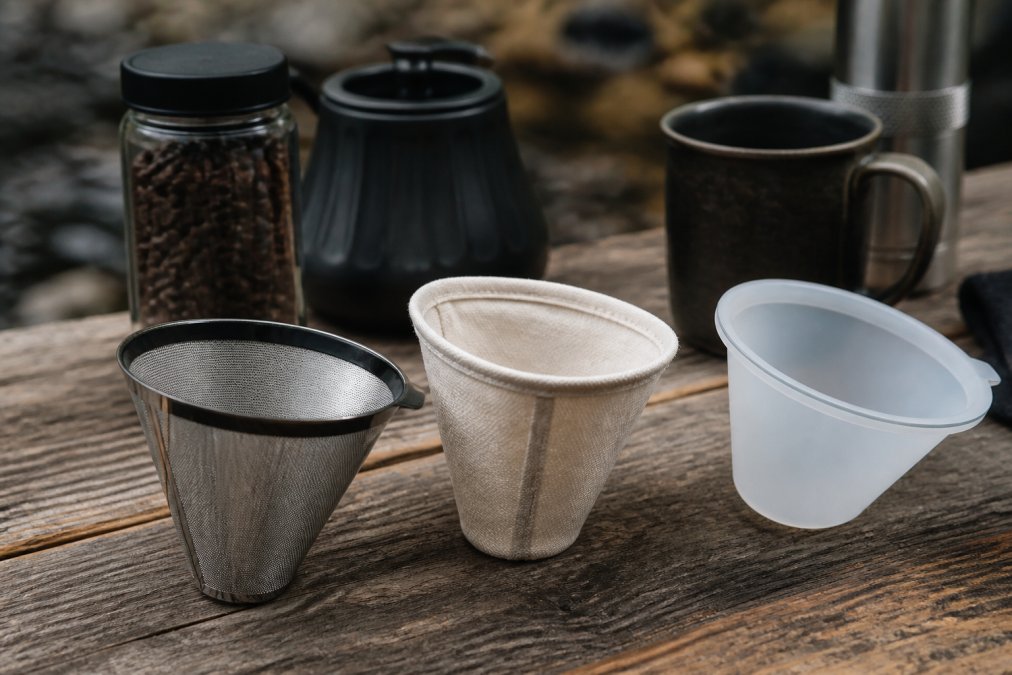 Tætbillede af tre forskellige kaffefiltre (rustfrit stål, bomuld og silikone) lagt side om side på en træbordplade med kaffe og rejseudstyr i baggrunden
