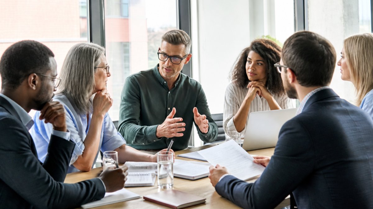 Diverse team of professionals in a modern meeting room collaborating at a table with notebooks and laptops, natural lighting from large windows, women and men engaged in discussion