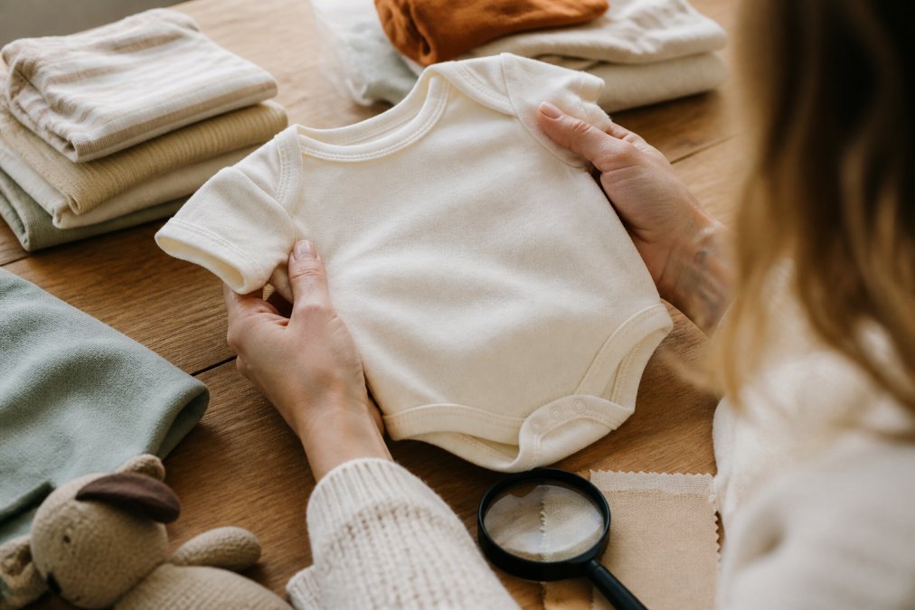A parent's hands holding a soft cotton baby onesie next to a magnifying glass and a fabric swatch, warm natural light on a wooden table