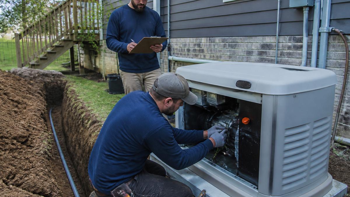 A licensed electrician in a residential backyard connecting wiring to a newly placed standby generator while a second worker reviews a clipboard, with a house and open trench visible in the background
