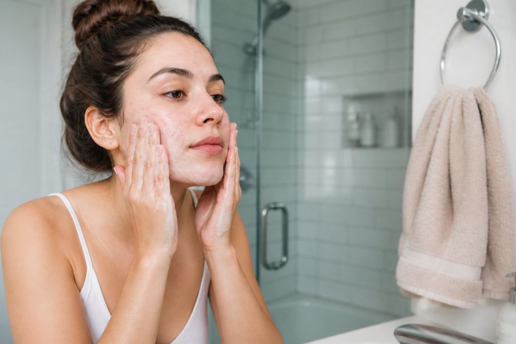A woman with acne-prone skin gently massaging a creamy, low-lather cleanser onto her damp face in a bright, clean bathroom, with a soft facial towel nearby