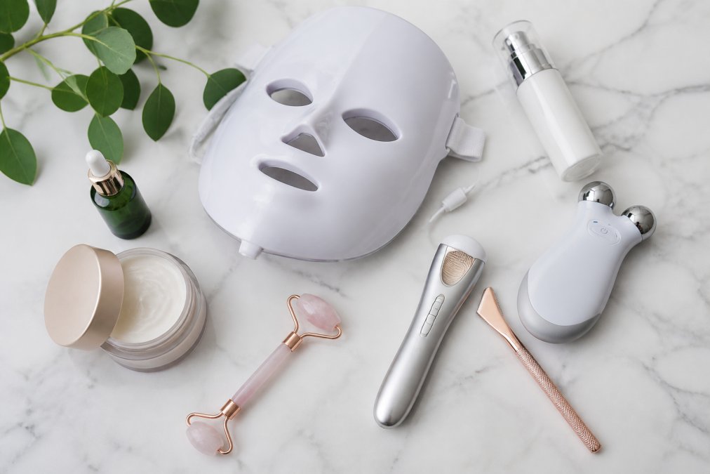 Flat lay arrangement of professional-grade at-home skincare devices including microcurrent facial tools, LED masks, and dermaplaning tools displayed on clean white marble surface with skincare serums and moisturizers