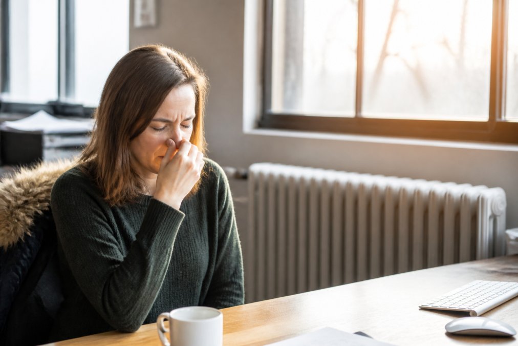 Person in a heated office environment touching their nose with visible discomfort, dry winter sunlight streaming through windows, showing the effects of low humidity on nasal passages
