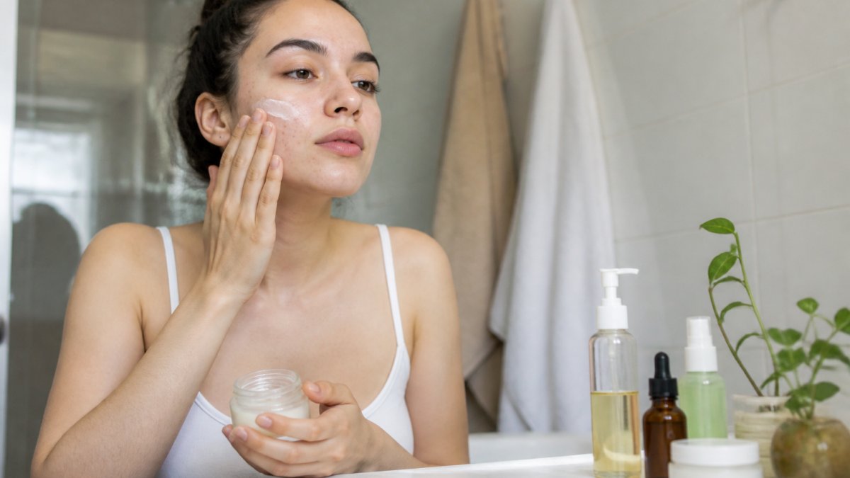 A young woman with sensitive, acne-prone skin gently applying moisturizer in a bright bathroom, with a simple lineup of skincare bottles on the counter