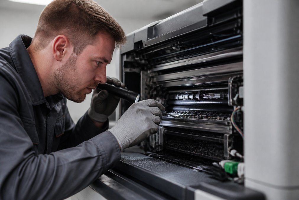 Maintenance technician inspecting the interior of a professional laser printer, carefully examining printheads and internal components with precision tools and lighting
