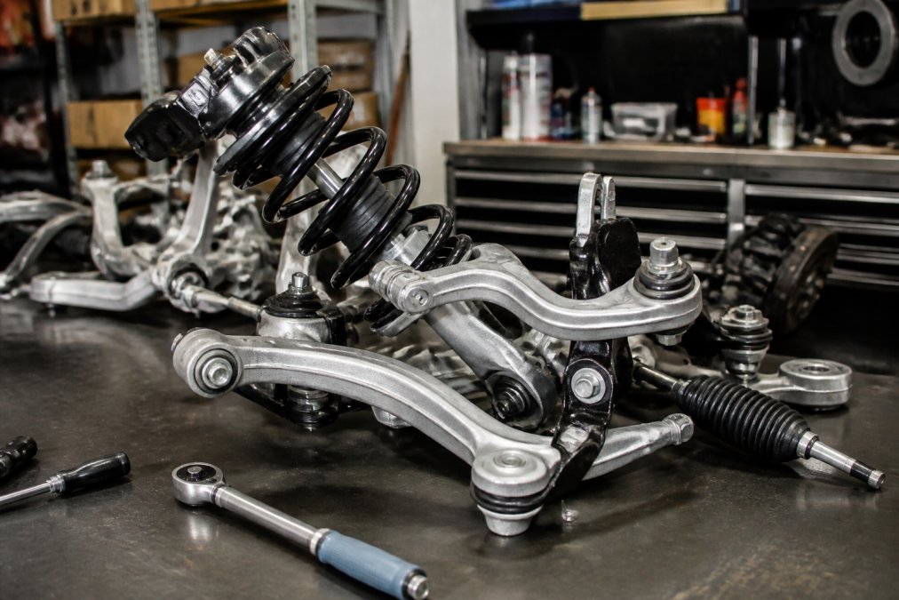 Close-up of a Porsche 911 suspension assembly showing struts, control arms, and ball joints mounted on a workbench with precision tools and diagnostic equipment nearby