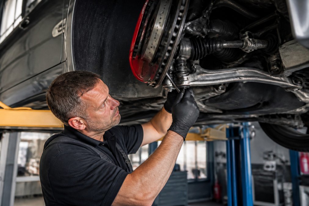 Experienced mechanic inspecting a vehicle's suspension system, checking control arms and ball joints with precision tools on a lift in a professional garage with bright overhead lighting