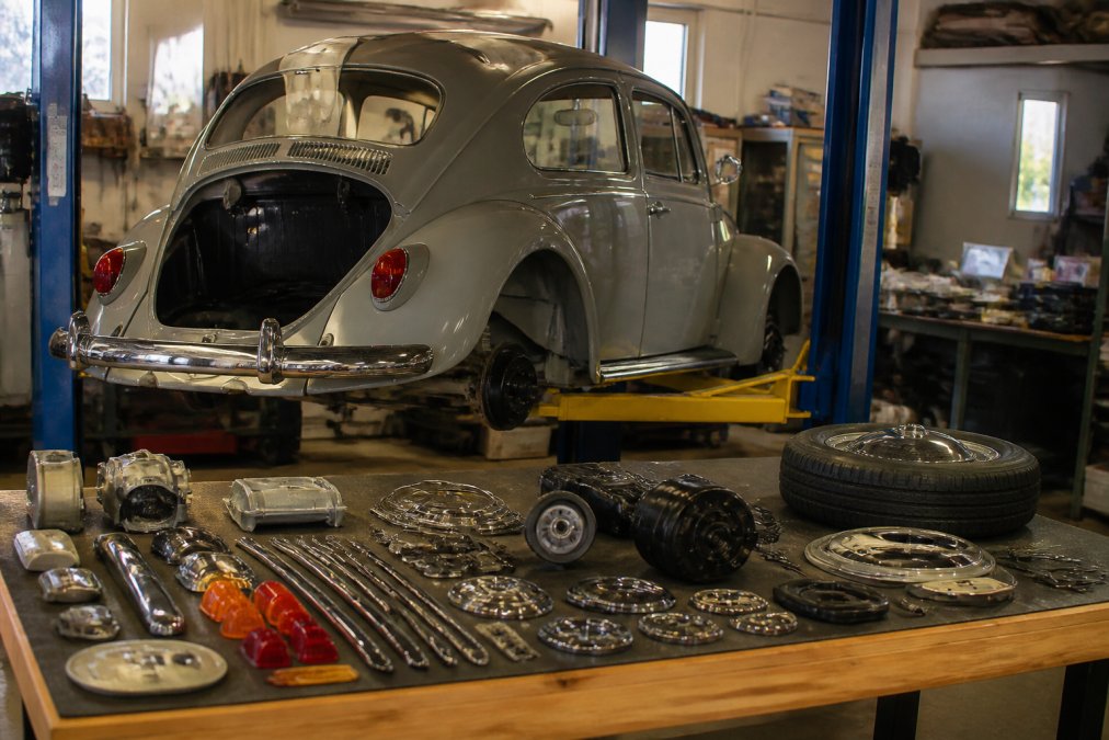 A classic Volkswagen Beetle in mid-restoration on a garage lift, with an organized array of genuine OEM parts laid out on a nearby worktable under bright overhead shop lighting