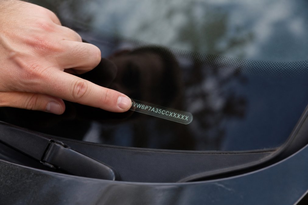 Close-up of a hand holding a vehicle identification number plate visible through a car windshield, with fingers pointing to the specific VIN characters for documentation