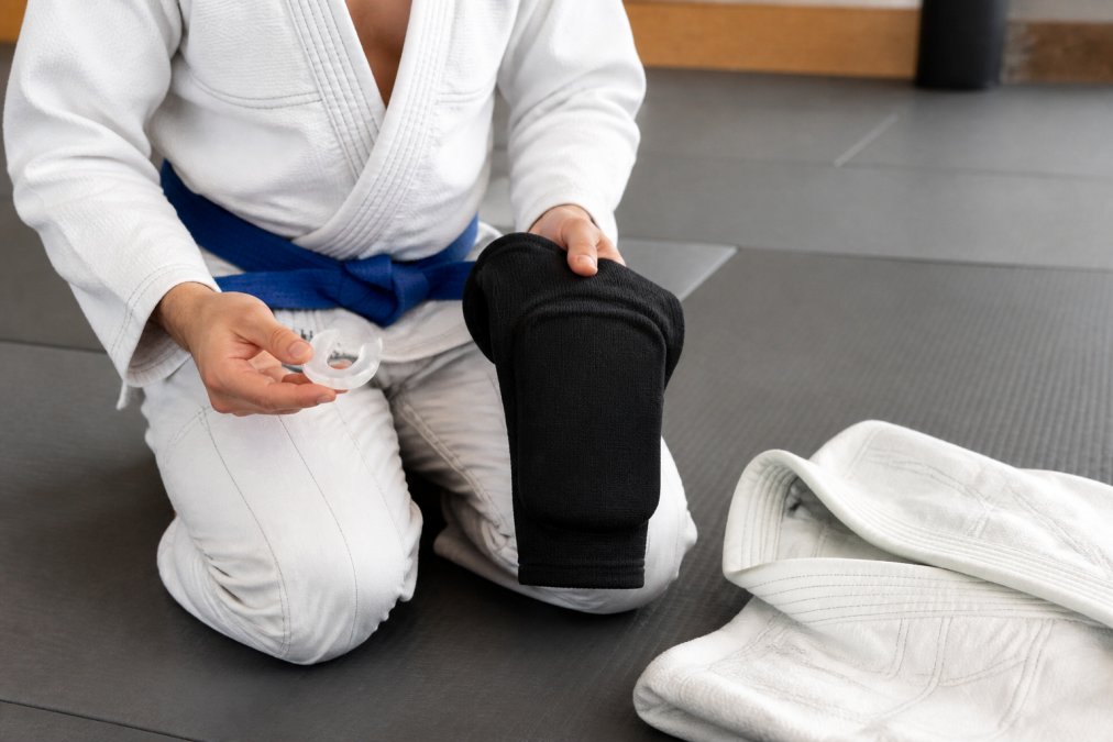 Close-up van een jujitsu beginner die kniebeschermers en een gebitsbeschermer vasthoudt naast een Gi op een tatami mat in een trainingsruimte