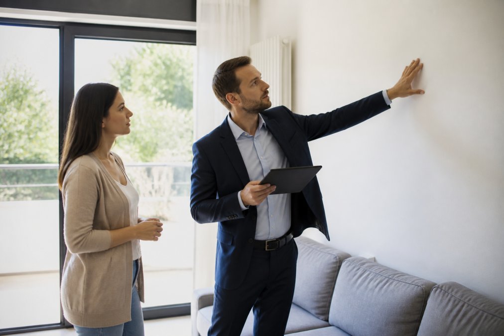 Gestionnaire immobilier professionnel examinant l'état d'un bien avec un locataire, inspectant les murs et les installations dans un salon lumineux avec éclairage naturel