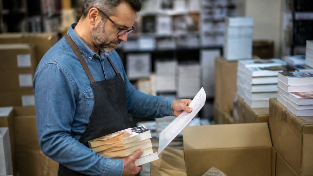 Un libraio che verifica i libri ricevuti da un fornitore, confrontando il documento di trasporto con i titoli e i codici ISBN sulla confezione, in un magazzino di libreria